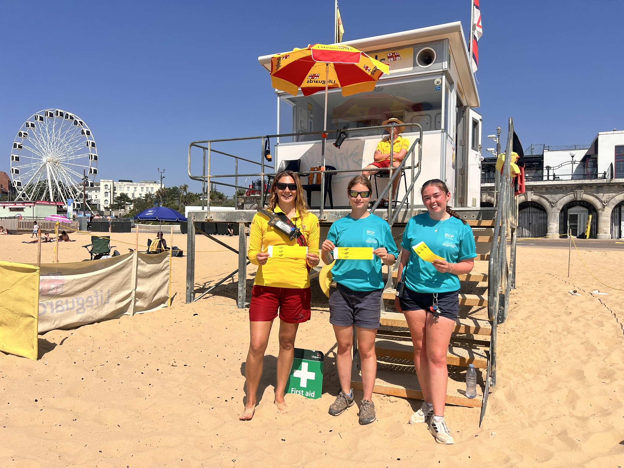 RNLI lifeguard and two KidZone staff members holding KidZone wristbands in front of an RNLI Lifeguard tower on Bournemouth Beach